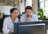 Two office workers at a shared desk look alarmed at a monitor displaying a full-screen alert.