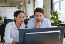 Two office workers at a shared desk look alarmed at a monitor displaying a full-screen alert.