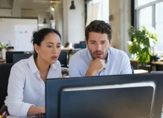 Two office workers at a shared desk look alarmed at a monitor displaying a full-screen alert.