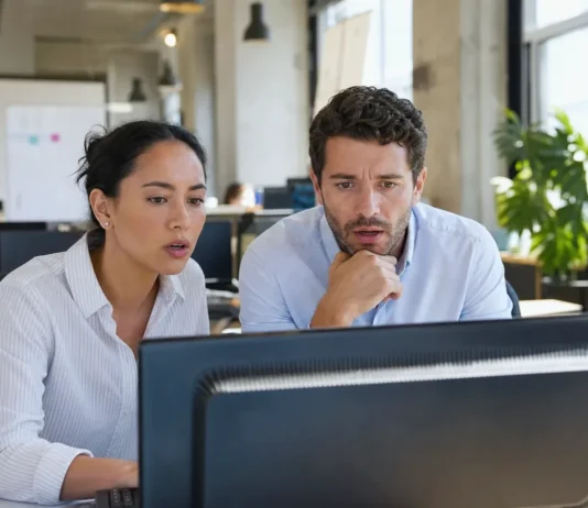 Two office workers at a shared desk look alarmed at a monitor displaying a full-screen alert.
