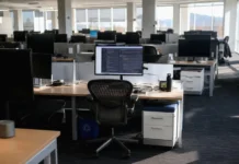 AI Security in 2027: 54% of Firms Can’t Limit AI Agent Access Brightly lit open-plan office with rows of desks and monitors; one active monitor at a central workstation.