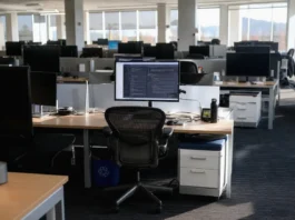 AI Security in 2027: 54% of Firms Can’t Limit AI Agent Access Brightly lit open-plan office with rows of desks and monitors; one active monitor at a central workstation.