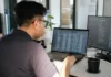 A person with glasses sits at a desk, focused on a laptop displaying data, in a bright modern office.