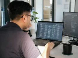 A person with glasses sits at a desk, focused on a laptop displaying data, in a bright modern office.
