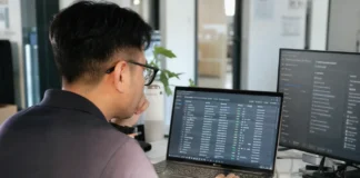 A person with glasses sits at a desk, focused on a laptop displaying data, in a bright modern office.