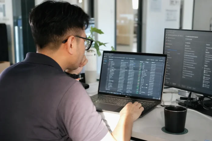 A person with glasses sits at a desk, focused on a laptop displaying data, in a bright modern office. A person with glasses sits at a desk, focused on a laptop displaying data, in a bright modern office.