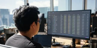 A software engineer sits at a desk in a bright open-plan office, working on a large monitor displaying data.