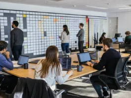 A spacious office with a long table of laptops, people working