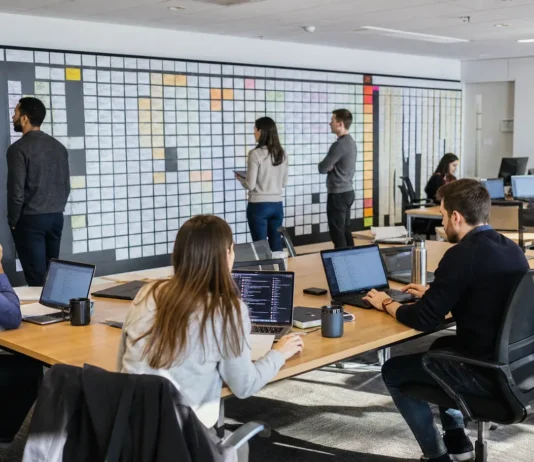 A spacious office with a long table of laptops, people working