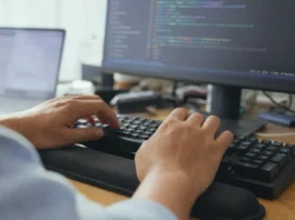Close-up of hands mid-keystroke on a keyboard, with a blurred monitor displaying code in the background.