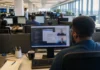 A modern open-plan office with rows of desks and monitors, brightly lit, featuring a person at a workstation.