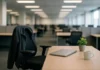 An empty corporate office workstation with a chair, jacket, laptop, and plant, illuminated by overhead lights.