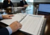 CISO Personal Liability Fears Nearly Double as AI Governance Mandates Expand Close-up of two hands on an open governance binder with dense, lightly defocused pages on a boardroom table.