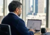 A man in a dark jacket sits at a desk, focused on a laptop screen in a bright office with city views.