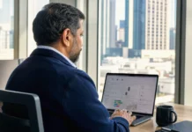 CSC CISO Survey: AI Security Optimism Runs Ahead of Domain Controls A man in a dark jacket sits at a desk, focused on a laptop screen in a bright office with city views.