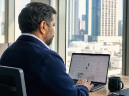 CSC CISO Survey: AI Security Optimism Runs Ahead of Domain Controls A man in a dark jacket sits at a desk, focused on a laptop screen in a bright office with city views.