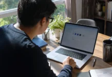 A person leans toward a laptop on a wooden desk, with a plant and books nearby, in natural light.