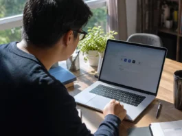 A person leans toward a laptop on a wooden desk, with a plant and books nearby, in natural light.