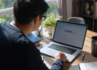 A person leans toward a laptop on a wooden desk, with a plant and books nearby, in natural light.