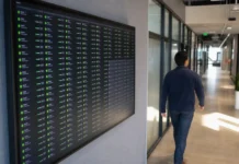 Enterprises Are Scrapping Agentic AI Pilots but Leaving Them Logged In A person walks past a large wall-mounted display showing a cloud-monitoring console with active AI agent sessions.