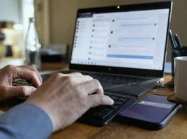 Close-up of hands typing on a keyboard, with a laptop screen displaying a chat panel and a smartphone nearby.