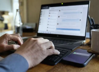Close-up of hands typing on a keyboard, with a laptop screen displaying a chat panel and a smartphone nearby.
