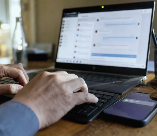Your MFA Won’t See This: Identity Security’s Blind Spot Inside Teams and Slack Close-up of hands typing on a keyboard, with a laptop screen displaying a chat panel and a smartphone nearby.