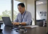 Healthcare Ransomware Attacks Shift: Businesses Up 35% While Hospitals Hold Flat Clinic administrator working at a laptop in a bright medical office, stethoscope on the desk and an examination room visible through the doorway
